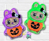 Two plush toys in Halloween-themed costumes on a white brick background. One is purple with a skull bow, the other green with a jack-o'-lantern face.